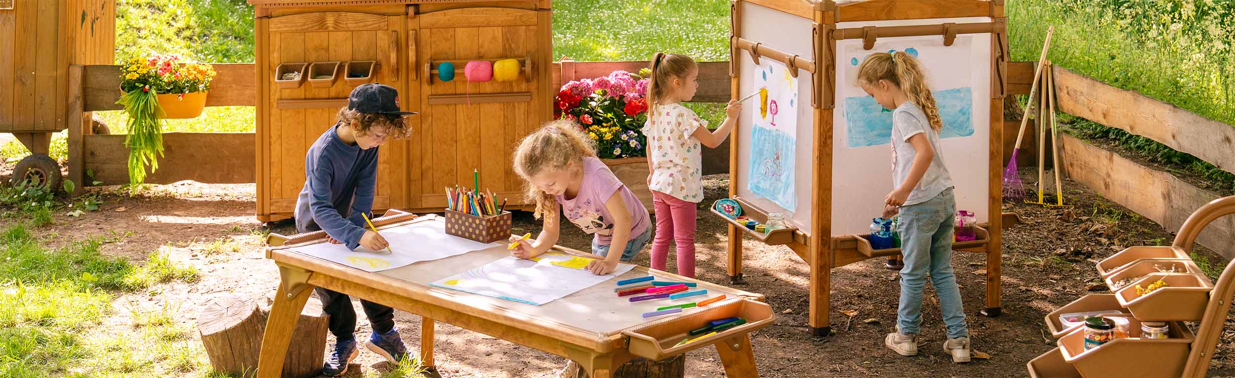 Ein Außenklassenzimmer für Vorschule und Grundschule, in dem Kinder an einem Außentisch und einer Staffelei lernen. Materialien werden in einem Kunstschuppen und auf einem Wagen aufbewahrt.