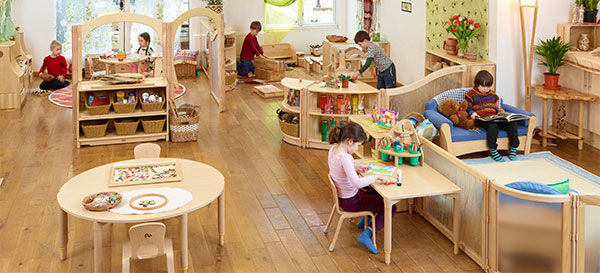 Ein Gruppenraum in einer Kita, wo Material in mobilen, flexiblen Regalen aufbewahrt wird. Kinder spielen auf dem Boden oder sitzen auf Stühlen und Sofas.