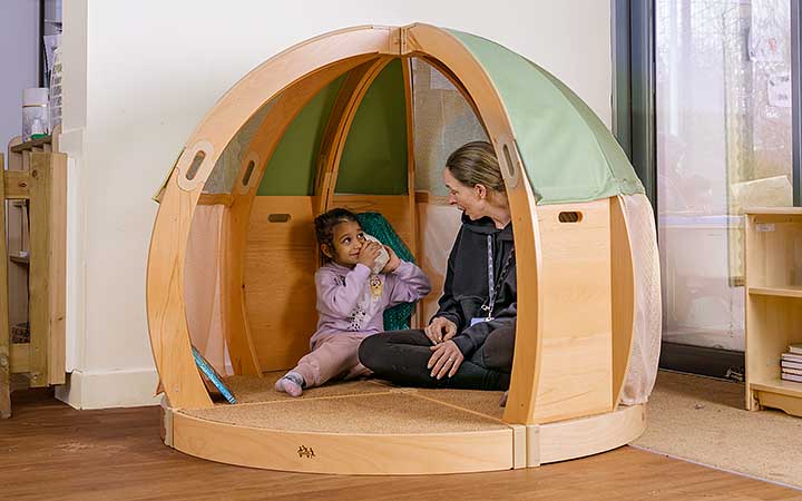 Ein Mädchen im Kindergartenalter und seine Bezugsperson erkunden gemeinsam eine Muschel in einer Spielhöhle aus Massivholz oder einem Spielbereich.