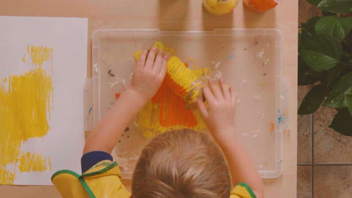 Kind rollt ein mit Schnur umwickeltes Papprohr auf einem Plastiktablett mit Farbe
