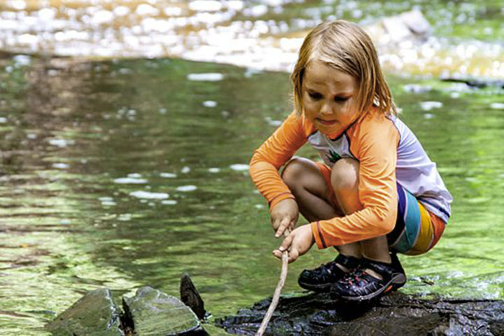 Ein Mädchen im Vorschulalter hockt auf einem Felsen in einem kleinen Bach und hält einen Stock