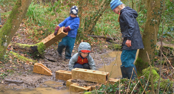 Drei Kinder in Regenkleidung bauen an einem Bach einen Damm aus Outlast Bausteinen