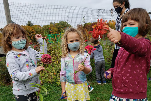 Kinder mit Masken im Garten halten Blumen, eine Erzieherin ist im Hintergrund