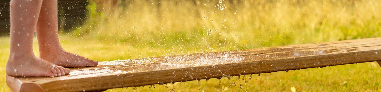 Nackte Kinderfüße stehen auf einem nassen Holzbrett, Wasser spritzt ringsum.