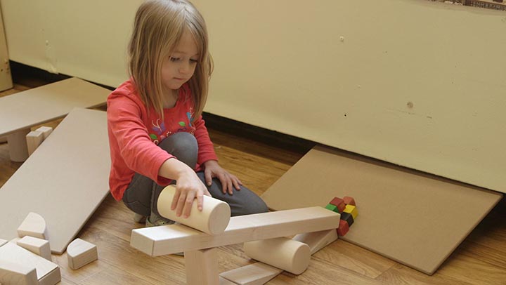 Ein Mädchen lässt einen Holzzylinder eine aus Holzbausteinen gebaute schiefe Ebene hinunter rollen
