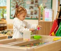 Girl in white sweater standing at a sand and water table and doing sensory play with water and small balls
