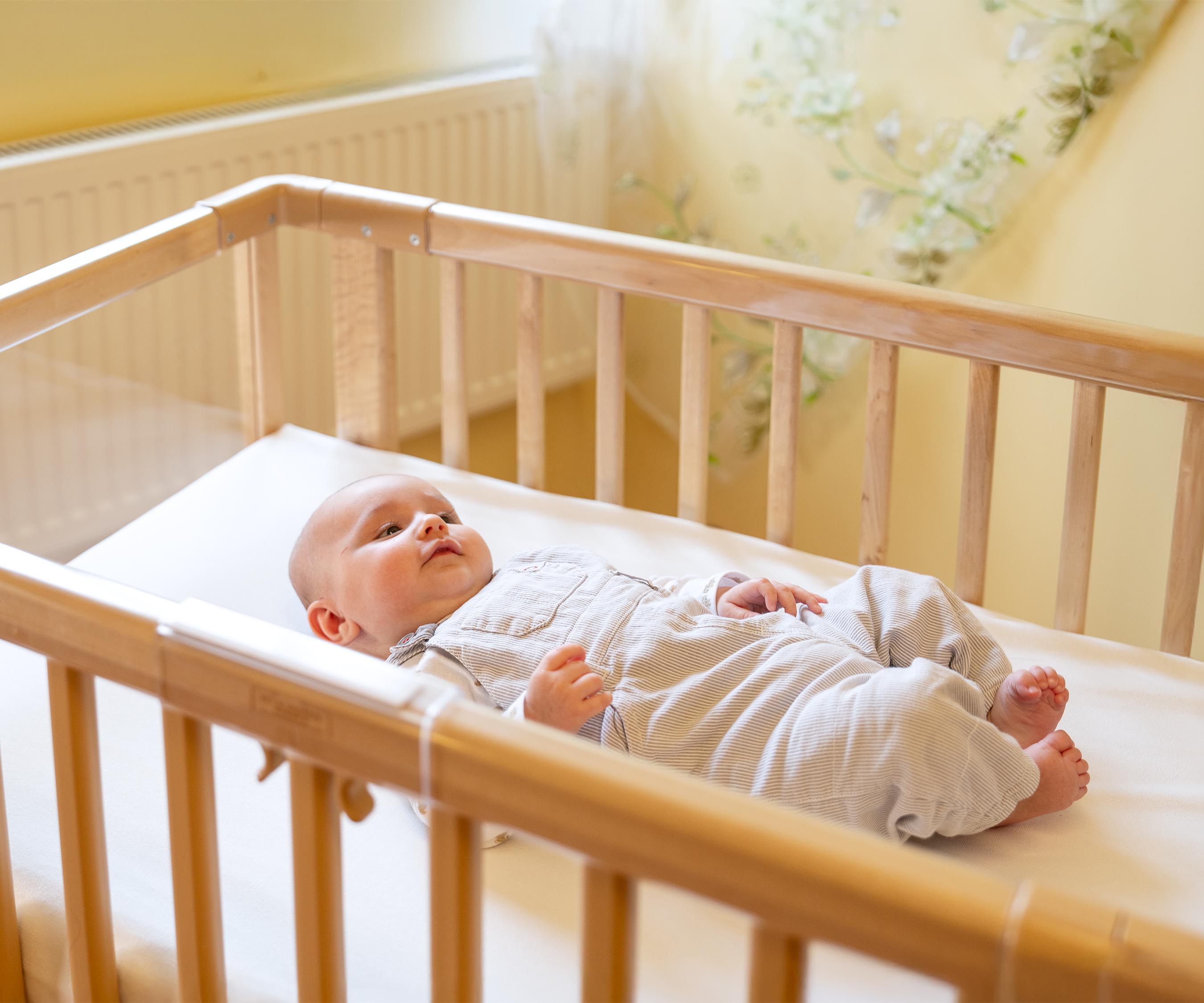 An infant with a grey romper and white shirt lying on hid back in a solid wood nursery cot.