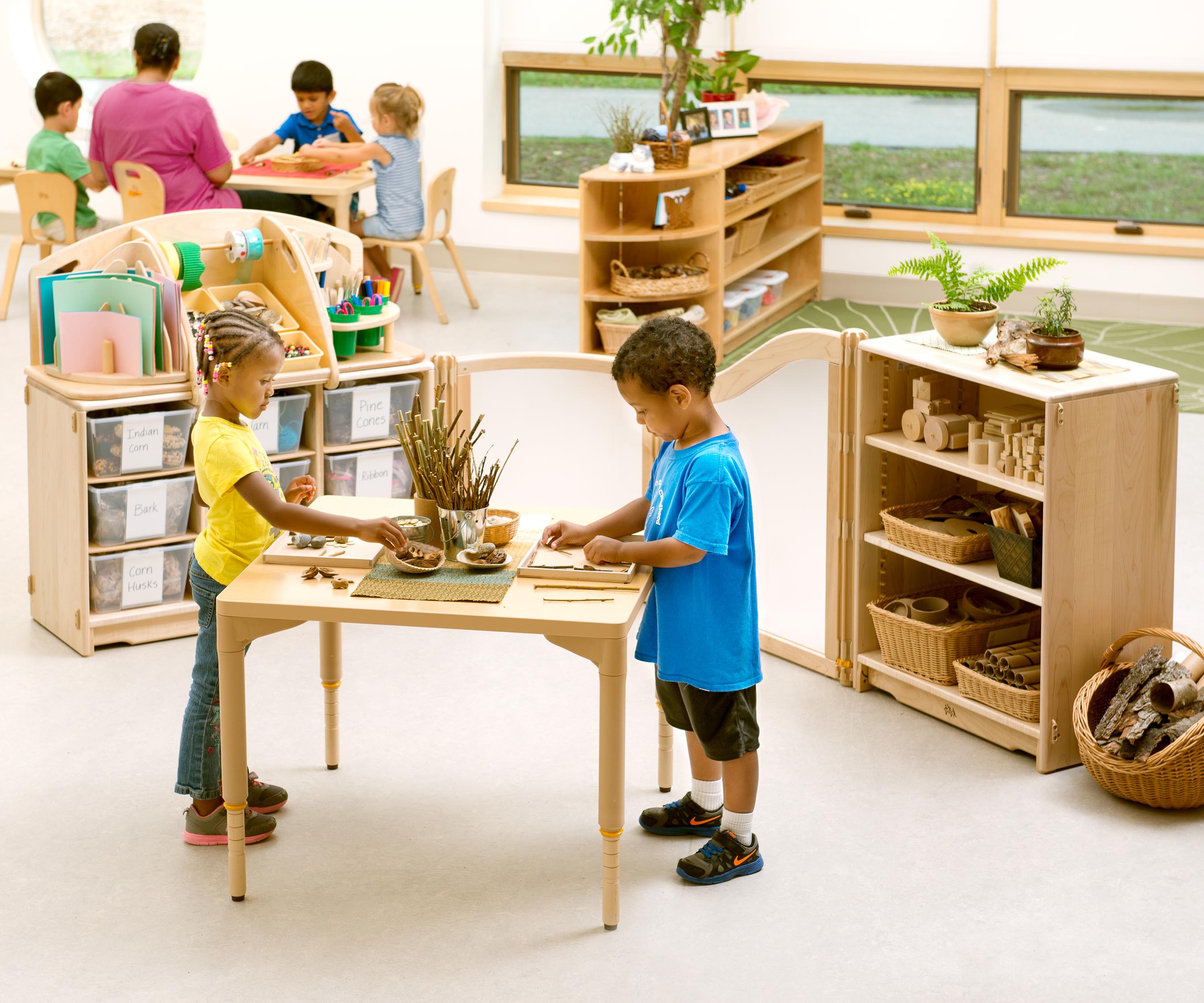 Zwei Kinder stehen an einem Tisch und beschäftigen sich mit Naturmaterialien, während im Hintergrund weitere Kinder und eine Erwachsene an einem Tisch sitzen.