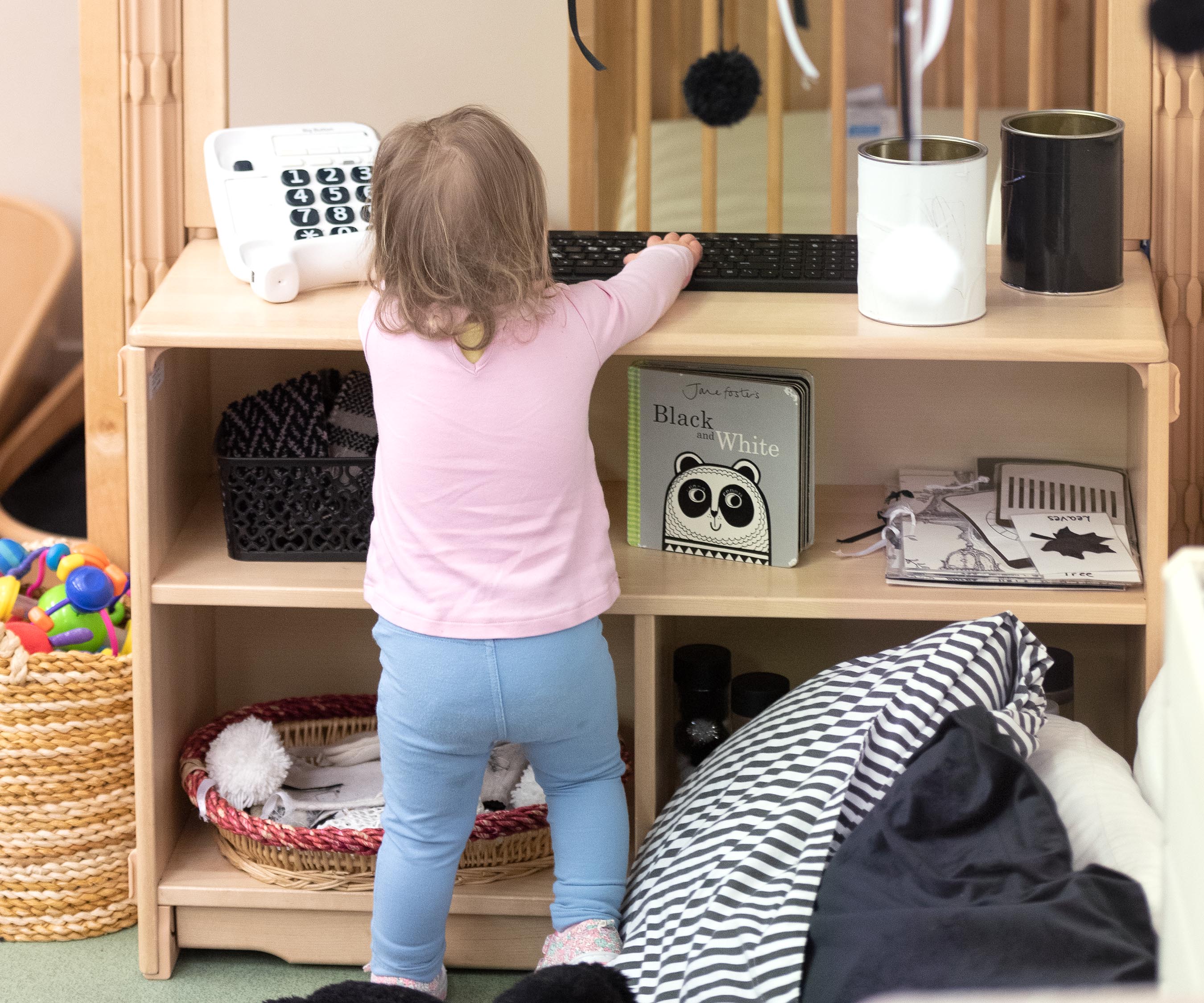 Baby dressed in pink and blue reaching for toy on solid wood nursery shelf
