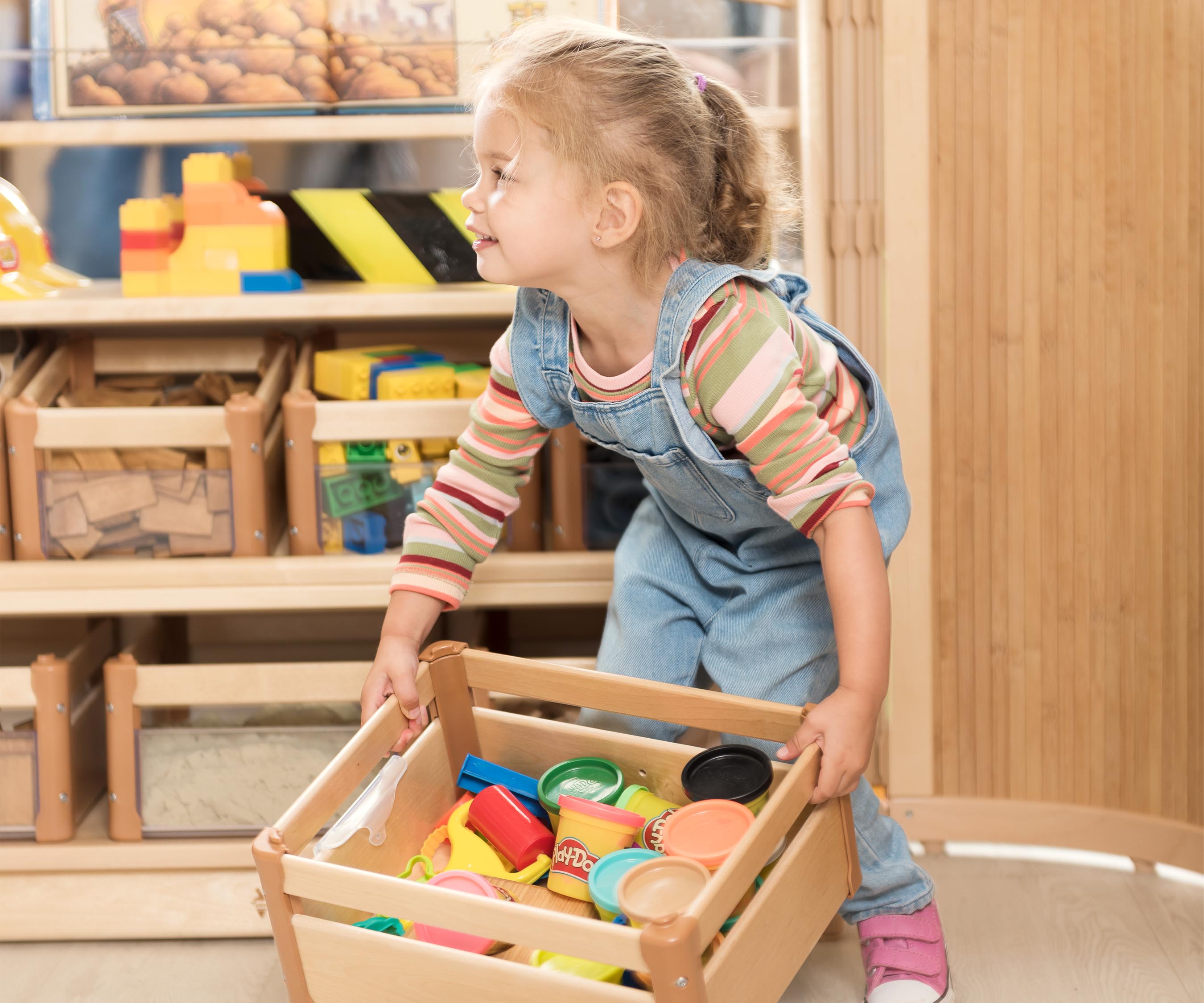 Mädchen in blauem Overall hebt einen mit Spielzeug gefüllten Holzkasten auf