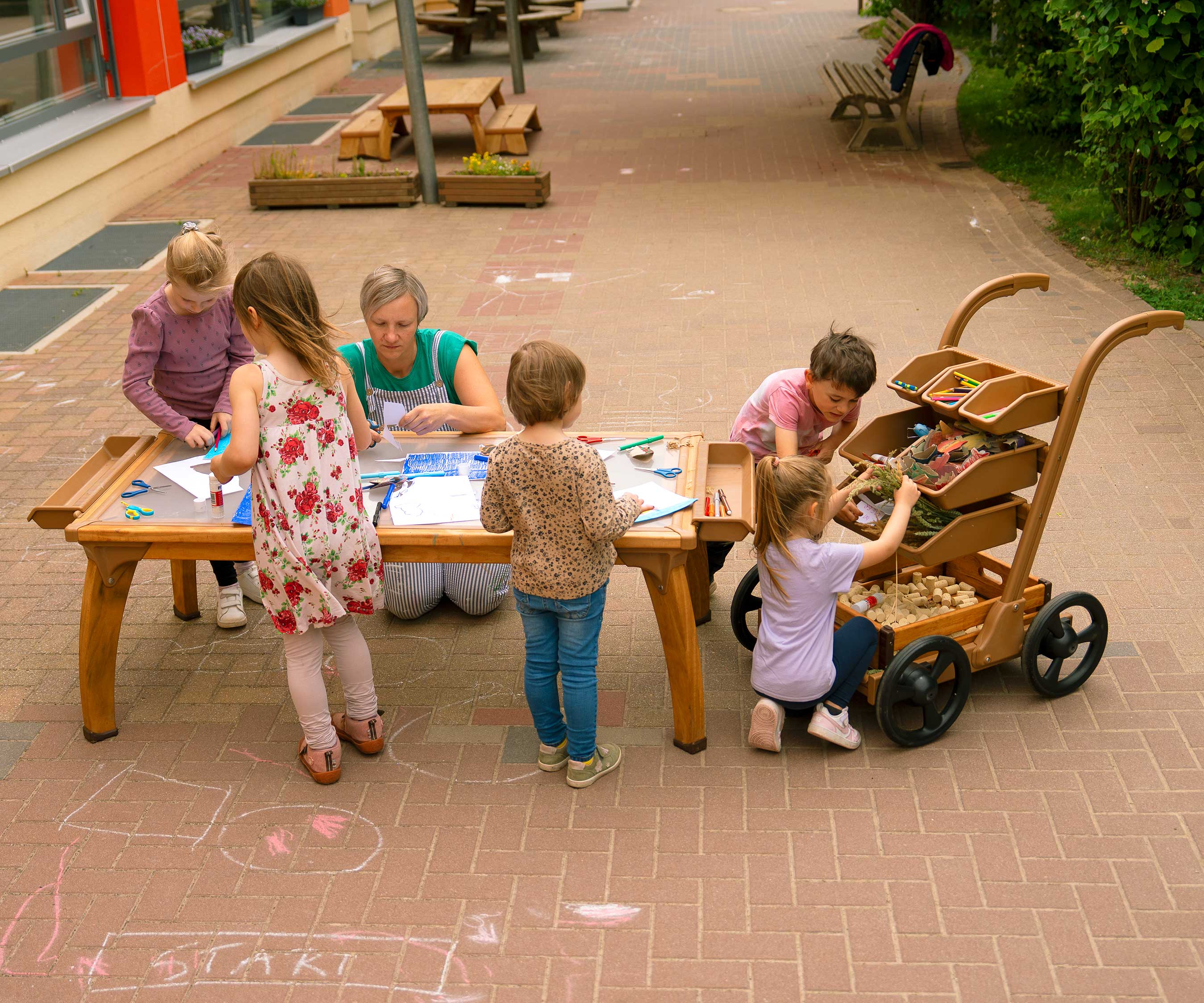 Kinder arbeiten in einem Outdoor-Kunstbereich mit einem Projekttisch und einem Wagen zum Aufbewahren und Transportieren von Materialien.