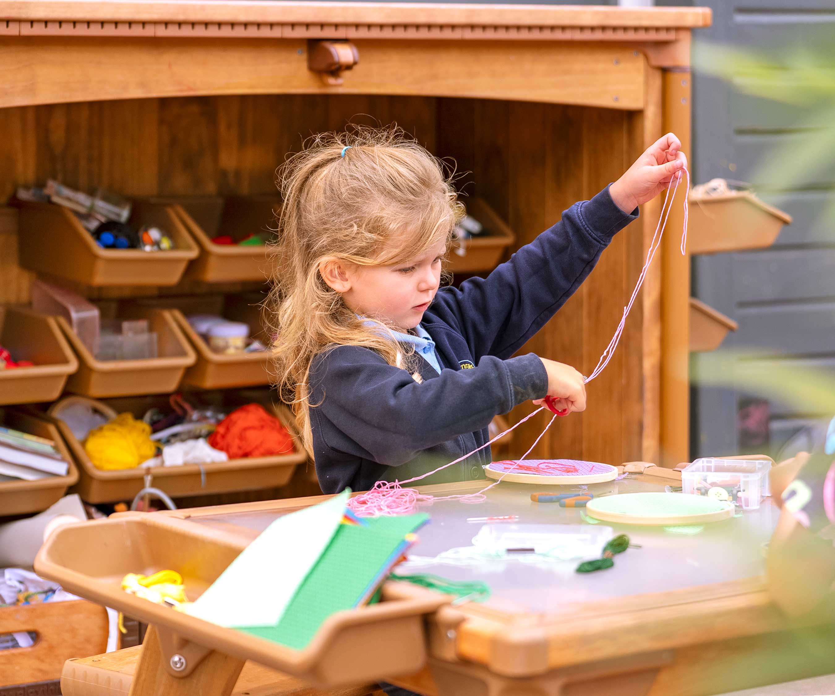 Ein Mädchen in einem Freiluftklassenzimmer einer Grundschule, das eine Nähaktivität durchführt, mit einem Kunstlagerhaus im Hintergrund.