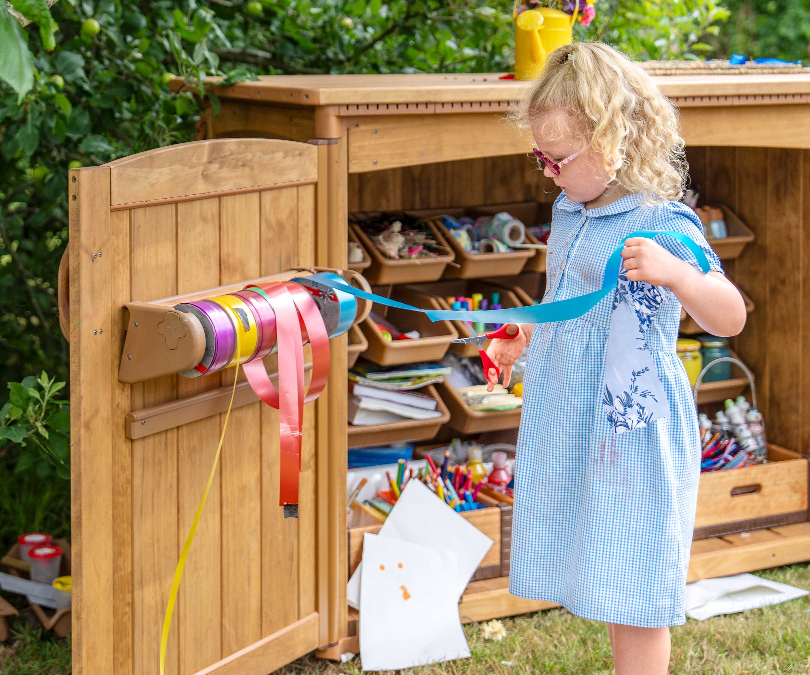 Ein Mädchen in einer Grundschulumgebung im Freien steht neben einem Kunstmaterialschuppen und schneidet ein Band von einem Halter ab.