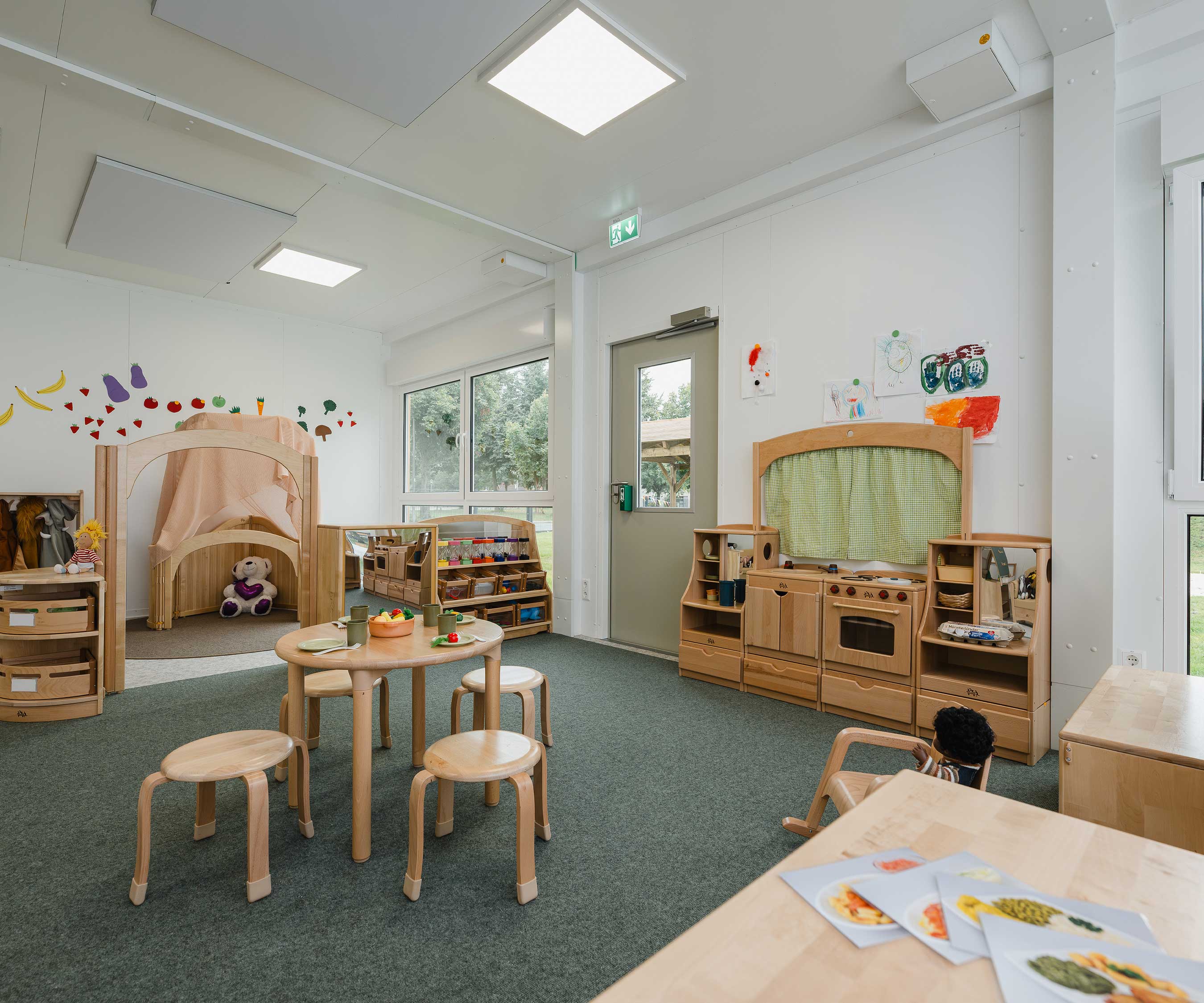 Ein Rollenspielbereich f&amp;#252;r eine Kita, der mit Massivholzm&amp;#246;beln und Spielzeug ausgestattet ist, darunter eine Spielk&amp;#252;che, ein Spieltisch mit Hockern und ein Ruhebereich.